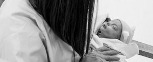 Una doctora sostiene a un bebé muy pequeñito. El bebé tiene los ojos cerrados y este enrollado en una cobija. Tiene un sombrerito de tejido