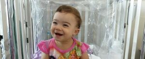 Una bebé ríe mientras está sentada en una cama de hospital. La cama está rodeada con y un plástico.