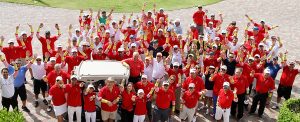 Grupo de personas numeroso, vestidas con camisas rojas y mangas de rallas blancas y rojas, saludan y sonríen a la cámara mientras posan para la foto.